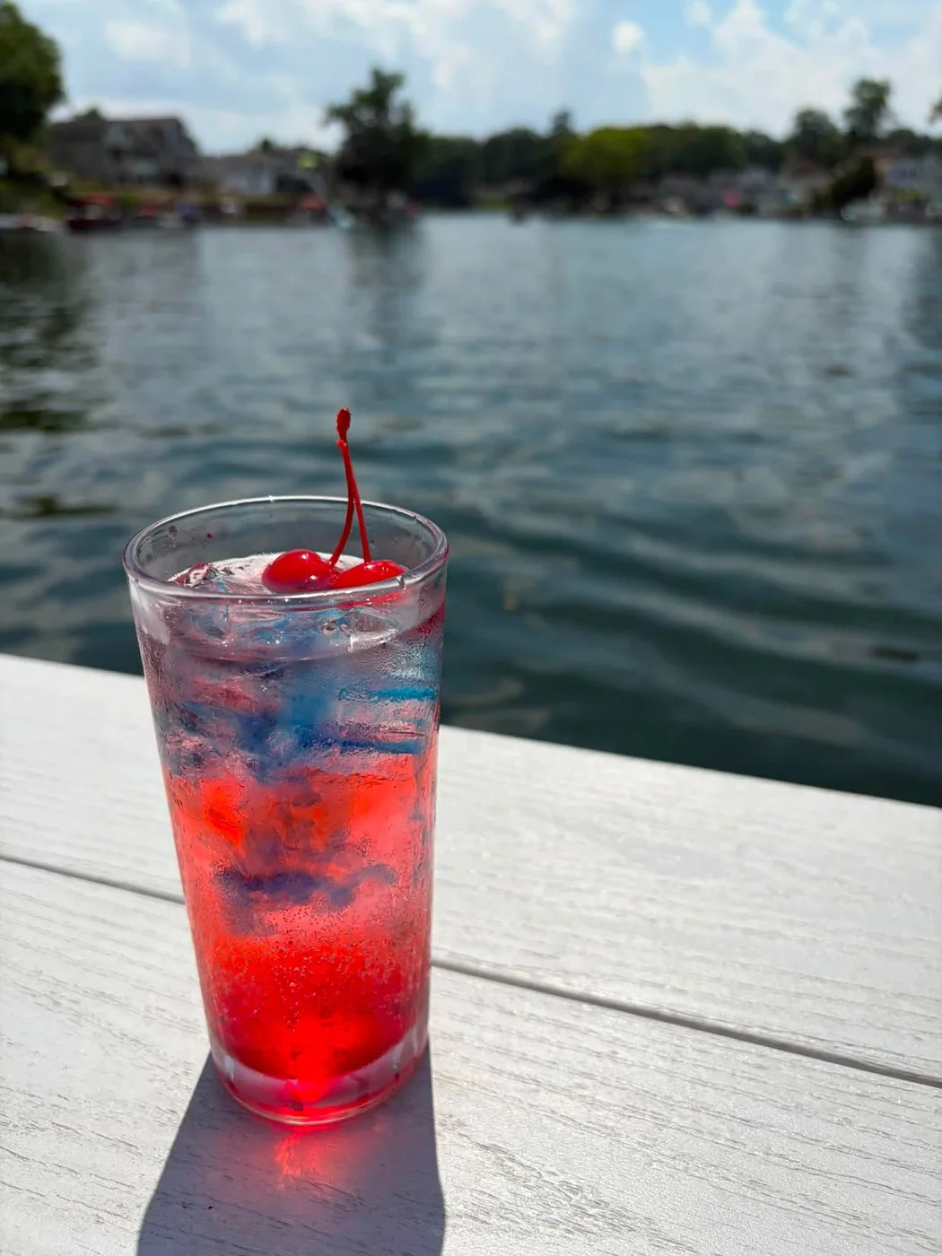 A tall red and blue layered cocktail with a cherry, on the white dock rail with the lake behind it