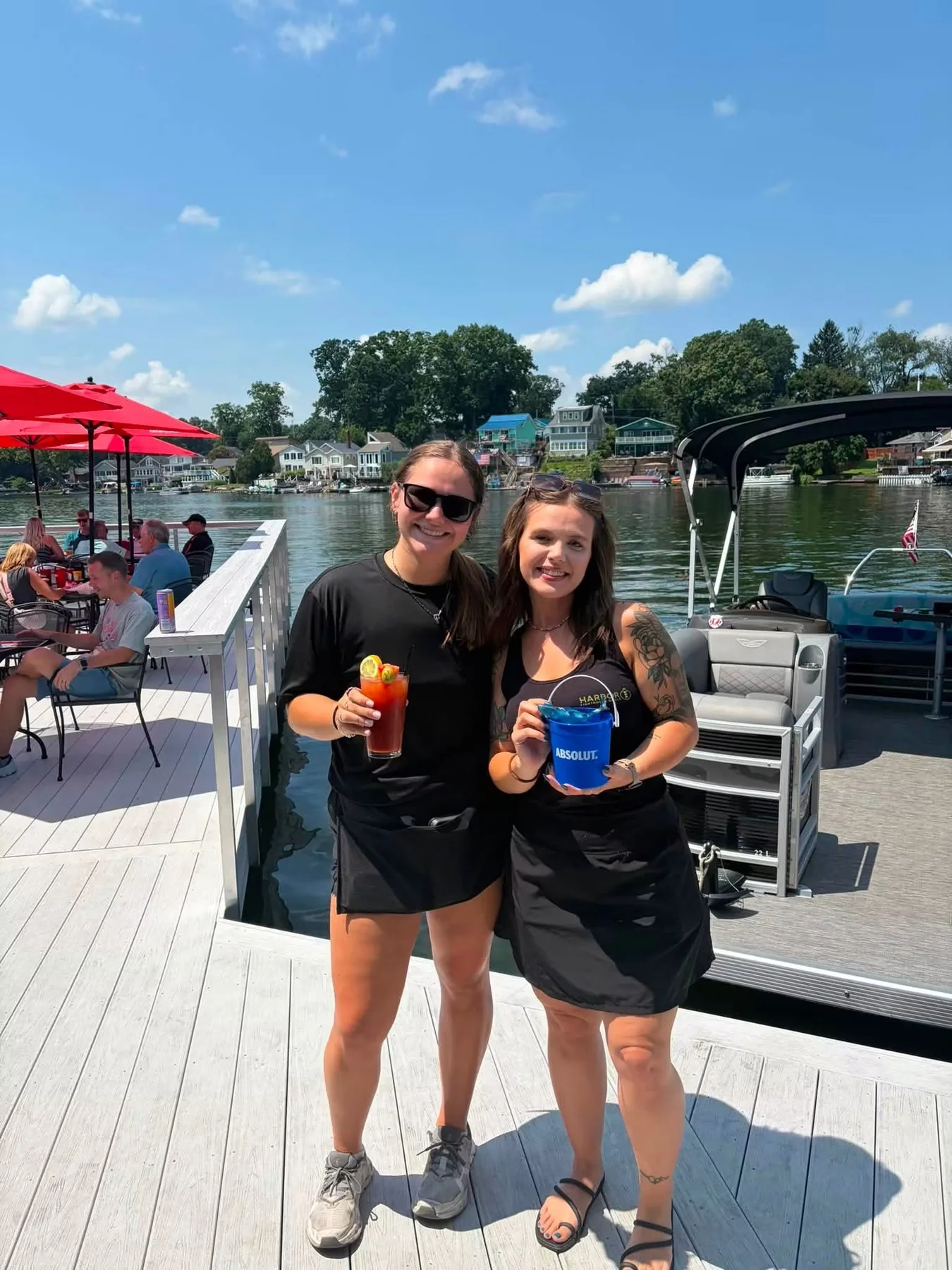 Two Harbor Lighthouse Grille servers on the deck over the water, one holding a cocktail, the lake and boats behind them